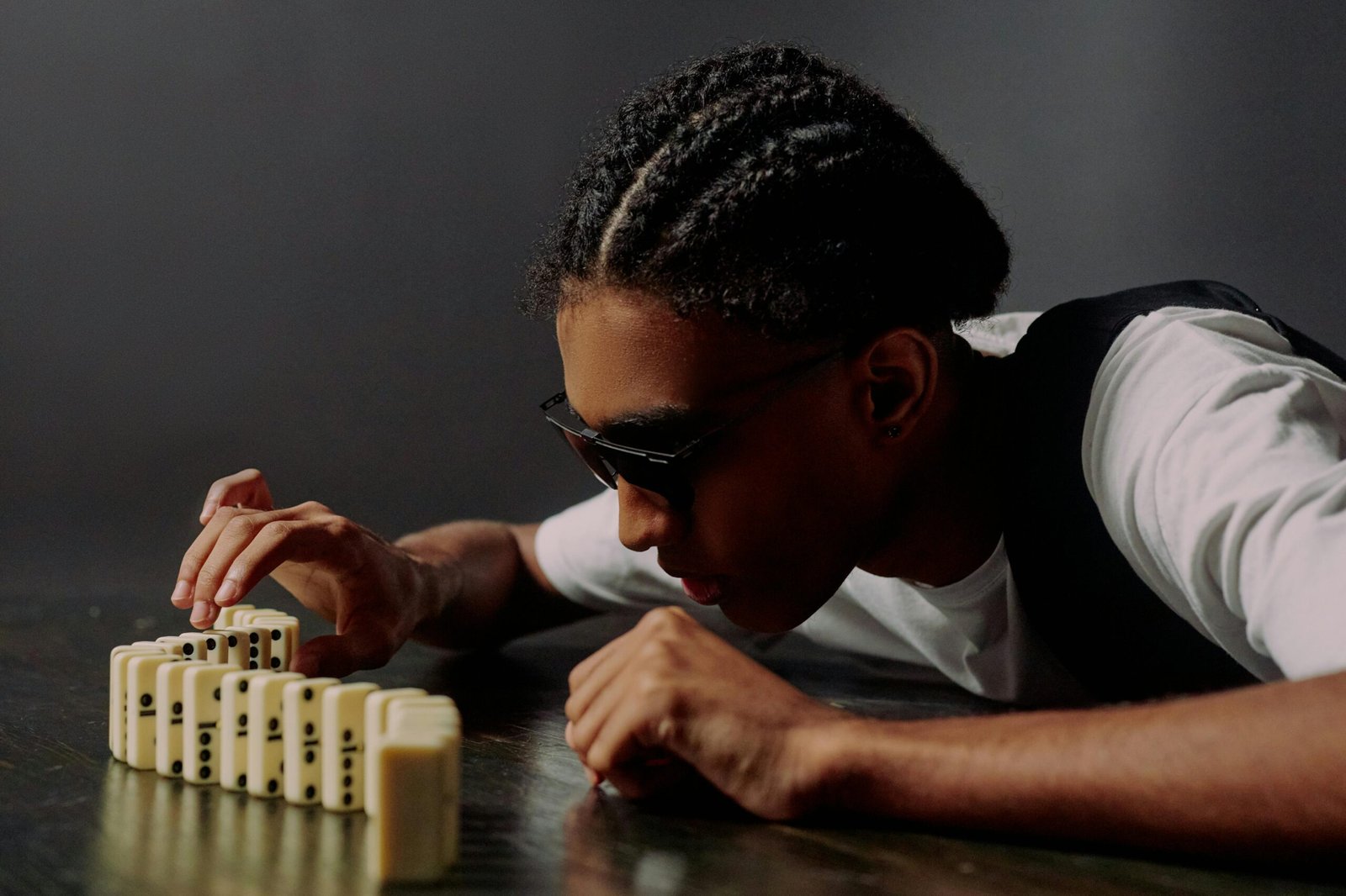 A man wearing sunglasses carefully lines up dominoes on a reflective surface, showing concentration and focus.