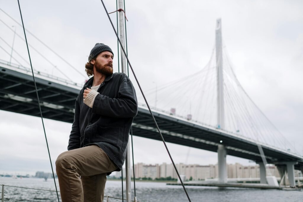 A man in a beanie on a boat near a large cable-stayed bridge, reflecting an adventurous journey.