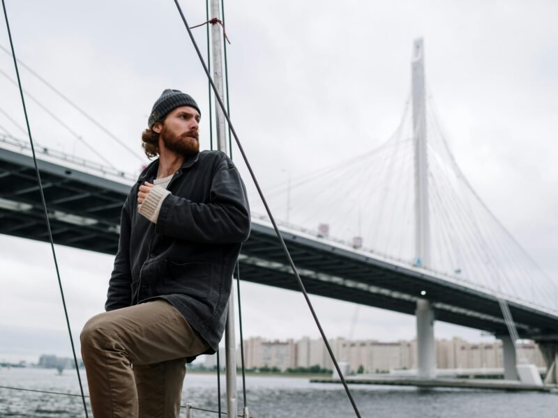 A man in a beanie on a boat near a large cable-stayed bridge, reflecting an adventurous journey.