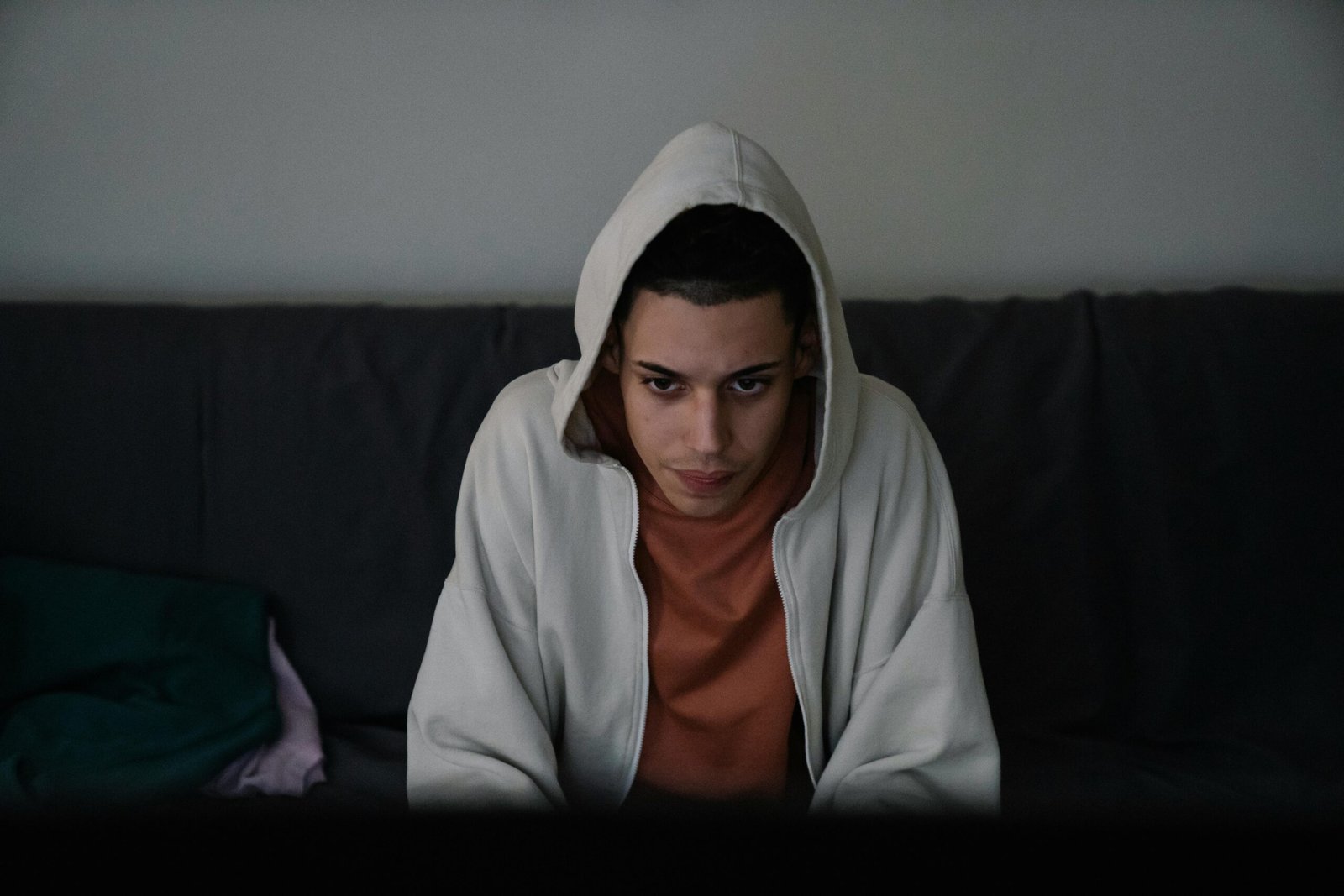 A young man wearing a hoodie sits on a couch, looking serious and concentrated inside a dimly lit room.