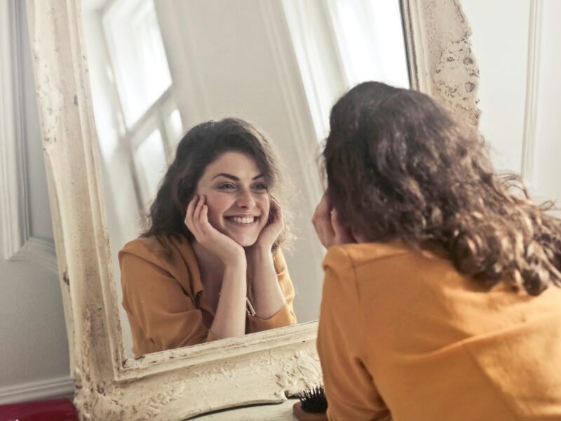 A cheerful woman smiles at her reflection in a vintage-style mirror, exuding positivity and warmth.