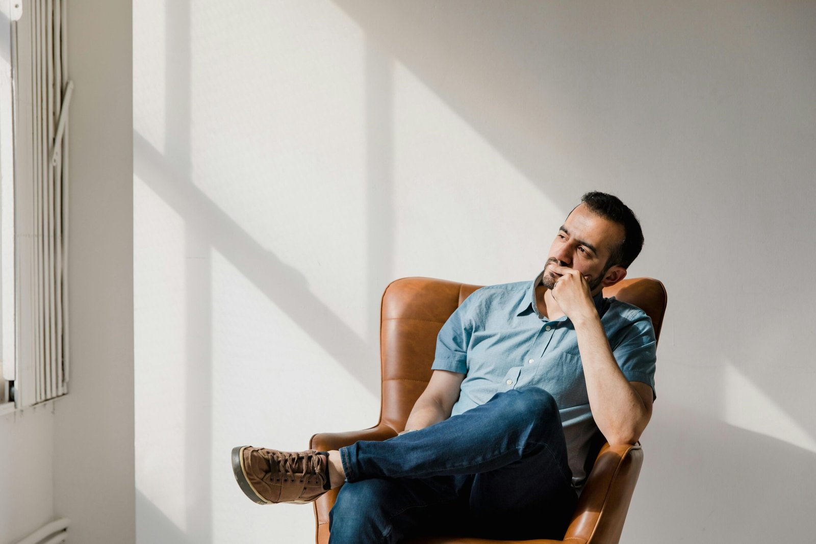 Man in casual attire sitting thoughtfully in a sunlit room, deep in contemplation.