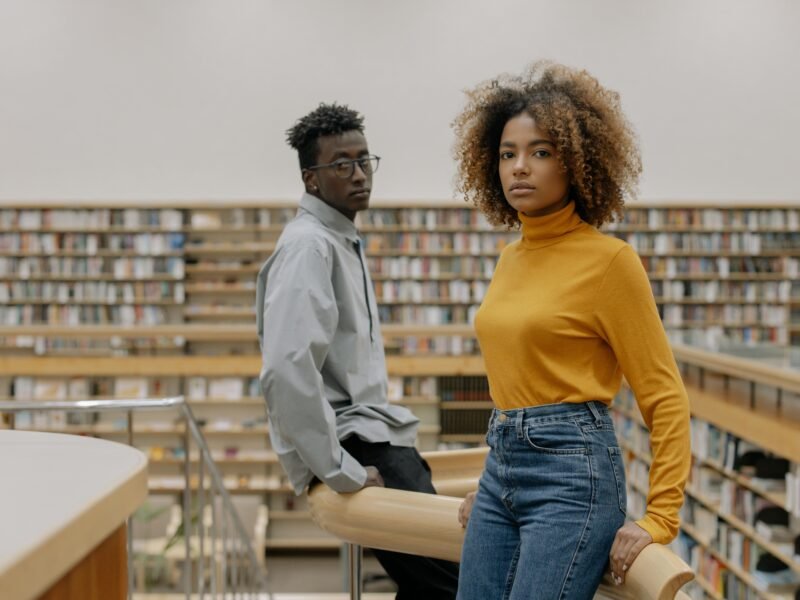 Two stylish adults pose confidently in a modern library with bookshelves in the background.