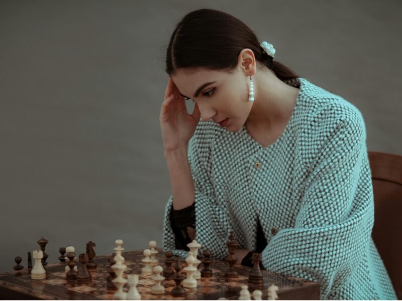 Concentrated ethnic female sitting on chair and touching head while playing chess against blurred gray background