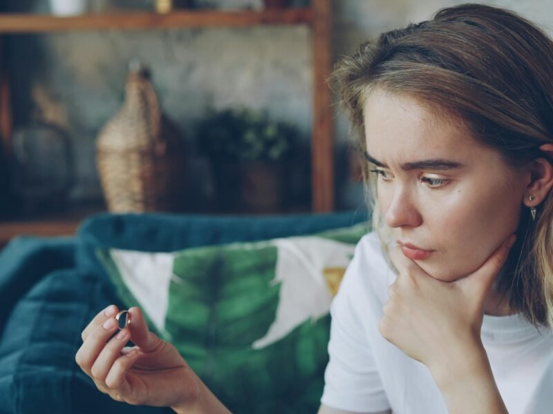 Young woman sitting indoors, holding a ring thoughtfully, looking pensive.