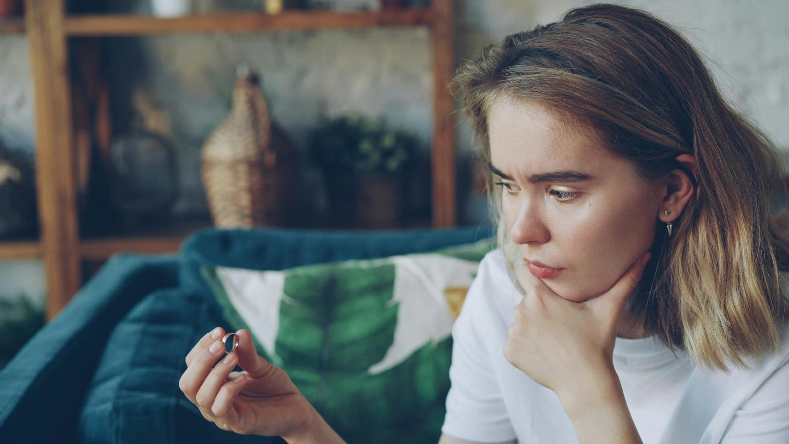 Young woman sitting indoors, holding a ring thoughtfully, looking pensive.