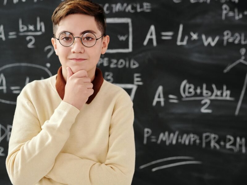 Smart student in glasses posing confidently in front of a blackboard full of mathematical equations.