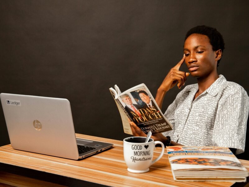 A young man reads a book at a desk with a laptop and mug, focusing thoughtfully.