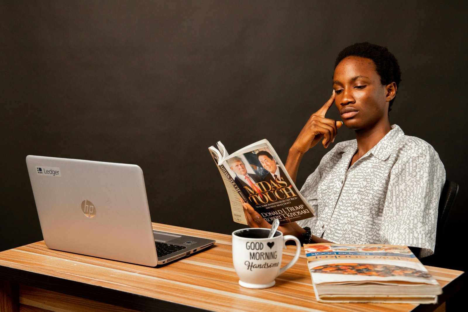 A young man reads a book at a desk with a laptop and mug, focusing thoughtfully.